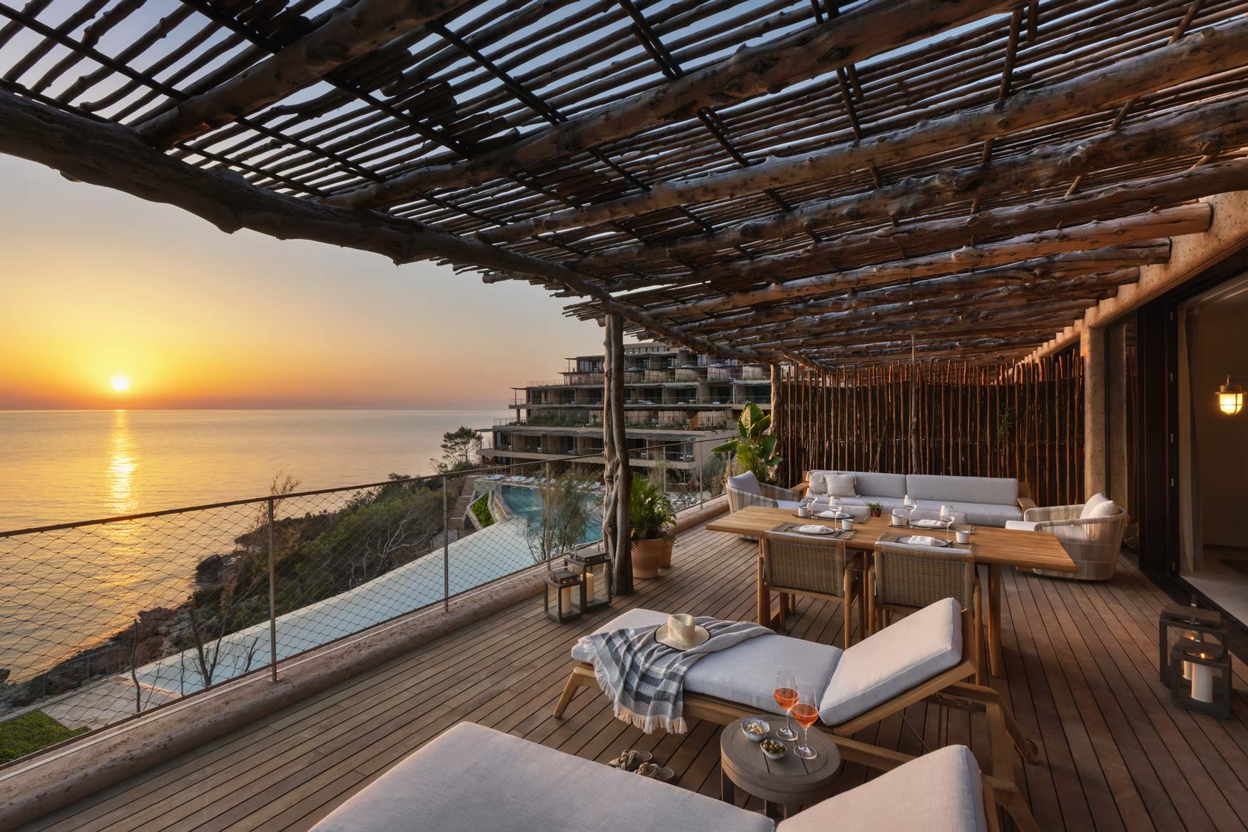 Terraza con pérgola de troncos sobre el acantilado de Cala Xarraca al atardecer, con el resort al fondo